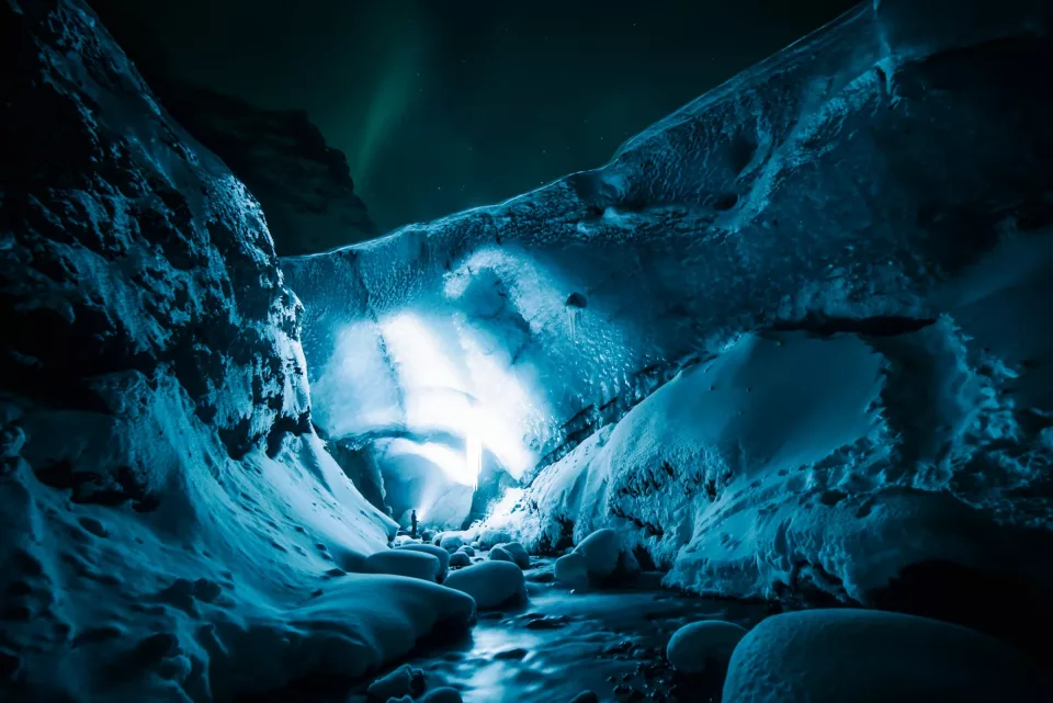 Une silhouette minuscule, lampe frontale allumée, au fond d’un canyon de glace bleue ; une aurore boréale verte dans le ciel de nuit
